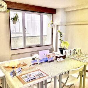 「天智開道」仙台鑑定会 B会場。大きな窓から柔らかい光が差し込む空間です。テーブルには薄い若草色のクロスが広がり、店主自身が撮影した写真をポストカードにして並べています。その隣には下からライトで照らされた水晶のフラーレンが置かれ、穏やかな光を放っています。窓辺のグリーンが生命の息吹を添え、落ち着いた色合いのインテリアが心を解きほぐす、自分自身と向き合い、未来の道筋を穏やかに見つめるための鑑定会場です。