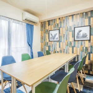 「天智開道」北千住C鑑定会の会場。木目調の明るい大きなテーブルを囲むように、ブルーやグリーンの鮮やかな椅子が並ぶ、お洒落なカフェのような雰囲気の部屋です。
奥の壁にはカラフルなウッドタイルのような壁紙と絵画が飾られ、大きな窓からは柔らかな光が差し込んでいます。
堅苦しい緊張感を忘れさせてくれるようなポップさと、明るい開放感に満ちており、初めての方でも肩の力を抜いて、前向きな気持ちで相談ができる鑑定会場です。