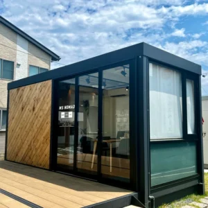 石屋蓮 仙台鑑定会を開催するコンテナ型レンタルスペースの外観。黒いフレームと斜めに張られた木目の外壁が特徴的なモダンなデザインです。正面は全面ガラスの引き戸になっており、室内には木製のテーブルや椅子、観葉植物が配置されているのが見えます。手前にはウッドデッキが広がり、青空が映り込む開放的な空間です。