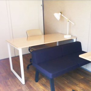 広島・西条鑑定会会場イメージ写真。モバイルスペース（Work）室内。明るい壁面で、部屋の奥に白い天板のデスクとデスクライト、奥側にキャスター付きの椅子が設置されています。手前側には紺色のソファ席が配置された個室空間です。
