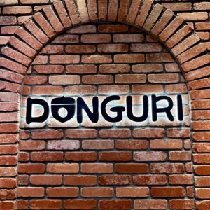 赤茶色のレンガ壁に、どんぐりのイラストと「DONGURI」の文字が白く光るバックライト看板。