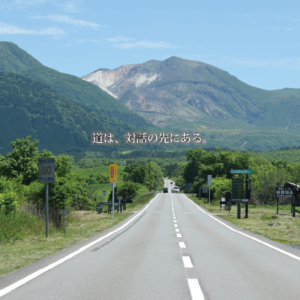 遥か彼方の山嶺へと続くまっすぐな道と、果てしなく広がる青空。道路標識と豊かな緑が並び、中央には「道は、対話の先にある。」という文字が置かれた風景。