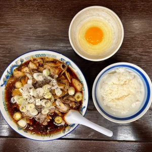 富山ブラックラーメンとライスのセットを上から撮影したもの。醤油の黒が際立つ濃厚なスープに、たっぷりのチャーシュー、太いメンマ、大ぶりの刻みネギと黒胡椒が容赦なく振られたラーメン。その傍らに白米と生卵の小鉢が並ぶ。