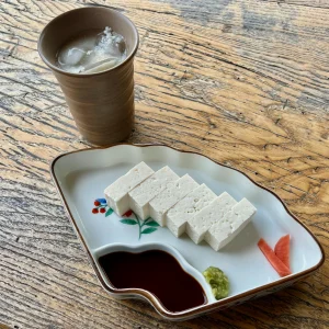 五箇山の相倉集落の店で提供された豆腐。扇型の美しい和食器に、ずっしりと密度の高そうな四角い白豆腐が並び、醤油、ワサビ、赤カブの漬物が添えられている。傍らには氷の浮かんだ酒の入った陶器のカップ。