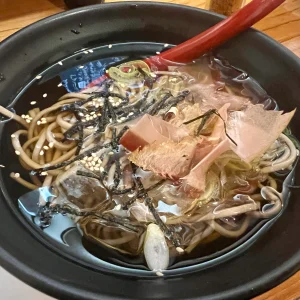 富山の居酒屋で提供された八尾の蕎麦。黒い器に盛られた蕎麦には透明な氷が浮かび、鰹節、刻み海苔、白ゴマ、ネギが贅沢に乗せられて、見るからに冷たくて喉越しが良そうな一杯。