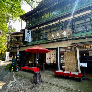 大岩山日石寺の参道沿いにある「大岩館」の店舗外観。新緑の木々の下に佇む歴史ある木造建築で、店先には赤い大きな野点傘と赤い毛氈が敷かれた長椅子が並び、風情ある景色を作り出している。