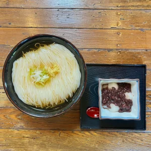 木目の美しいテーブルの上に並ぶ、大岩そうめんとぜんざいのセット。美しい黒い器に盛られたそうめんと、四角い小鉢に盛られたたっぷりの小豆のぜんざいが、黒いお盆に乗せられている。