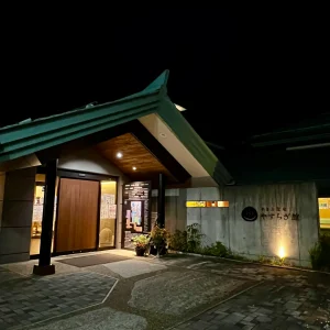 夜の闇の中に浮かび上がる「やまと温泉やすらぎ館」の入り口。特徴的な大きな三角屋根の車寄せがあり、温かみのある間接照明がコンクリートの壁と「やすらぎ館」の文字を静かに照らしている落ち着いた風景。
