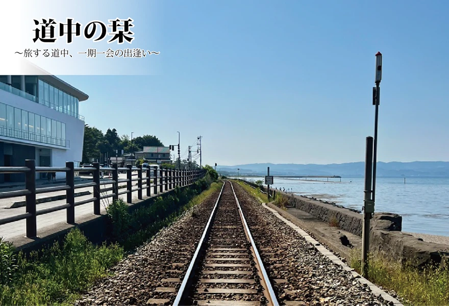 青く晴れ渡る空の下、穏やかな海沿いを真っ直ぐに伸びていく一本の線路の風景。左側には近代的な白い建物と柵があり、旅の途中の心地よい開放感が広がっています。左上には「道中の栞 〜旅する道中、一期一会の出逢い〜」という文字が配置されています。