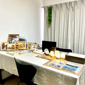 「天智開道」京都D鑑定会の会場。デスクには、自然な風合いが美しい薄茶色のリネンクロスが敷かれ、シックな黒い椅子が置かれた温かみのある部屋です。
クロスの上には、ぬくもりを感じる複数の球体照明や、木製フレームに飾られた美しい絵画が丁寧に並べられ、カーテンにはさりげなく緑の植物がそっと添えられています。
古補の落ち着きを感じる上品な静けさがありながらも、自然素材のファブリックが優しく緊張を解きほぐしてくれる、素直な心で深く向き合える鑑定会場です。