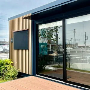 モダンな外観の鑑定会場。木製のルーバーと大きなガラス扉が特徴的で、店内の鑑定スペースがガラス越しに透けて見える。空模様はどんよりとしており、嵐の前の静けさを感じさせる。