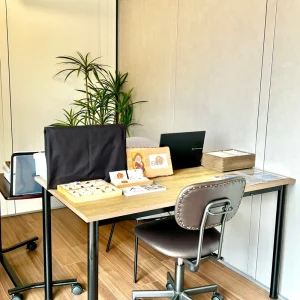 鑑定が行われていたデスク。木製のテーブルの上には店主のロゴが入った木製プレート、資料、そしてトレイに並べられた石が置かれている。背後には緑の植物が配置された、静かな空間。
