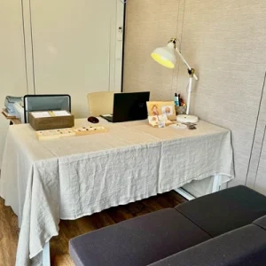 石屋蓮 広島・西条鑑定会の会場内。明るいベージュのテーブルクロスが掛けられた鑑定用デスクが置かれ、その上には温かな光を放つデスクライト、パソコン、石を並べた木箱、店主の看板が整然と配置されています。デスクの向こう側には椅子が備えられ、手前にはクライアント様用のグレーのソファが配置された、落ち着いた鑑定空間の様子。