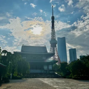 増上寺の歴史ある本堂の屋根と、その背後に高くそびえる東京タワー、さらに近代的なビル群が重なり合う東京らしい風景。空にはドラマチックに渦巻く雨上がりの雲が覆い、太陽の光が輪のように広がっている。