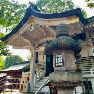 豊川稲荷東京別院の奥の院。端正な屋根の曲線と、手前に配置された重厚な石灯籠が調和している。柔らかな光が差し込み、建造物の細かな彫刻や木々の緑を幻想的に浮かび上がらせている様子。