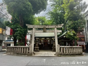 都会の恵比寿のビル街に囲まれた、石造りの鳥居が特徴的な神社の境内。手前には円状に組まれた石造りの玉垣があり、その中央に古びた石の鳥居がそびえ立つ。鳥居の奥には屋根が見え、境内の周囲は背の高い木々に覆われており、都会の喧騒から隔絶された独特の静寂と重厚感が漂っている。