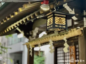 神社の境内に吊るされた、黒い木枠と組子細工が施された角形の灯籠。電球の明かりが灯籠の格子越しに温かく漏れ出ており、内部の紋章が薄っすらと浮かび上がる。背景には社殿の軒先と、湿った空気を纏う境内の木々が写り込み、夜の帳が下り始める前の、深く静かな社の中の情景を切り取っている。