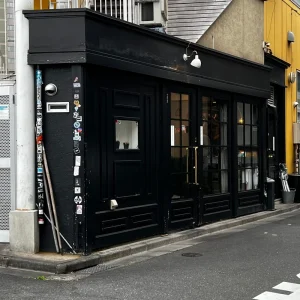 夕暮れ時の恵比寿にて、黒く塗られた重厚な扉を持つ店舗外観。壁面には無数のステッカーが貼られ、店舗備品と思われる棒が立てかけられている。静かな街角に佇む、店主が休息と鋭気を養うための隠れ家のような場所。