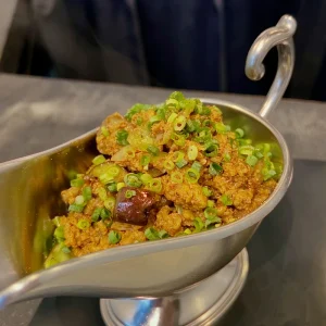金属製のグレービーボートに盛られた、スパイスと肉の荒々しい質感が伝わるキーマカレー。彩りに細かく刻まれた青ネギが散らされており、湯気と共にスパイスの強烈な香りが立ち昇ってくるような一枚。