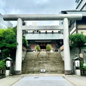 曇り空の下、芝大神宮の荘厳な石造りの鳥居が静かにそびえ立っています。中央の石段には夏越の大祓の茅の輪が設置されており、厳かな空気が漂っています。