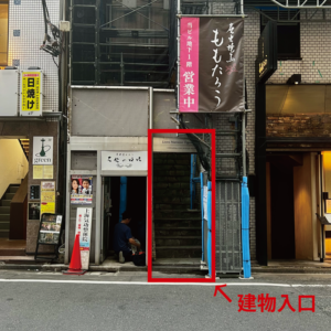 恵比寿鑑定会会場（ライオンズマンション小川恵比寿）の入り口写真。右隣に工事用の足場と青い緩衝材が設置されており、薄暗い階段へと続く入り口は赤い枠と矢印、そして「建物入口」のテキストで強調されています。周囲には「ももたろう」や「日焼け」などの看板があります。