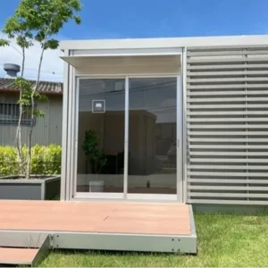 石屋蓮 奈良鑑定会会場となる、コンテナ型レンタルスペースの外観。すっきりとしたフレームと軽やかなルーバーが調和した、開放感のあるモダンなデザインです。ガラスの引き戸の前には心地よいウッドデッキが広がり、若葉をつけた木々が柔らかな木漏れ日を落としています。日常の喧騒からふっと解き放たれ、自分自身の本質と対話するための、風通しの良い穏やかな空間です。