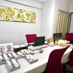 「天智開道」恵比寿C会場。壁面に飾られた植物のモチーフが描かれたアートが、空間に明るく華やかな彩りを添える鑑定会場です。白を基調とした清潔感のある室内には、深みのあるボルドー色のチェアが配され、凛とした落ち着きを感じさせます。デスクの上には、温かな光を放つ球体ライトや、丁寧に並べられた石のジュエリー、そして物語を紡ぐ御朱印帳が置かれ、訪れる方の心を解きほぐすような、穏やかな時間が流れる鑑定会場です。