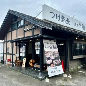 「つけ蕎麦 蕎麦令和」の外観。曇天の空の下、木の温もりを感じる店構えと、暖簾をくぐり抜ける旅の気配を切り取っています。
