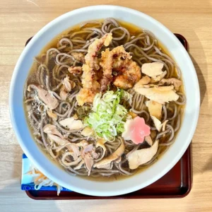 直径30センチメートルもの巨大な白い丼にたっぷりと盛られた、山形名物の「冷たい肉そば」特盛。透明感のある冷たい醤油ベースのつゆの中に、山のように盛られた蕎麦が沈んでいる。表面には親鶏のコリコリとした薄切りの鶏肉が散らされ、中央には鮮やかな緑色の刻みネギが小高く盛られている。さらに、蕎麦の上には揚げたての「げそ天」が香ばしげな衣を広げており、その横にはピンク色の花麩が一つ添えられている。丼は木製の盆に乗せられ、奥には木目のテーブルが見える。