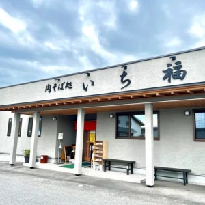 山形県東根市にある「肉そば処 いち福」の店舗外観。建物の壁はグレーを基調とし、軒下の木目がアクセントになっている。どんよりと厚い雲に覆われた空の下、店舗入口のガラス戸には赤い暖簾がかかっている。駐車場の奥には他の住宅や車も見えるが、人気は静まり返っている。ここで急遽検索し、素晴らしい味に出会えたことを想起させる風景。