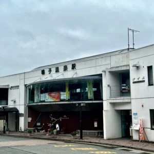 曇天の空の下に佇む、JR東日本の鳴子温泉駅の建物。白い壁に、丸みを帯びたガラス張りの特徴的なデザインの駅舎。駅正面には「鳴子温泉駅」と黒い文字で看板が掲げられている。駅前には少し人が歩いており、植栽のプランターも見える。周辺の山並みも遠景に薄く見え、これから始まる温泉街での湯治への期待感を醸し出している。