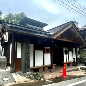 鳴子温泉にある共同浴場「古湯 滝乃湯」の入口付近。黒い瓦屋根と白壁、そして使い込まれた木の梁や柱が歴史を感じさせる、重厚な和風建築の佇まい。木の入口の前には暖簾がかかり、石碑には「古湯 滝乃湯」の文字が刻まれている。足元には小さな石灯籠や飛び石があり、建物からは温泉の湯気が立ち上って周囲の冷気と混ざり合っている。静かな温泉街の風情を伝える風景。