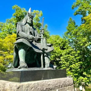 米沢・上杉神社の境内に佇む、青空を背にして力強く鎮座する謙信公の銅像。甲冑をまとい静かに刀を握るその姿は、周囲の豊かな緑と深い青空の下で、冷徹なまでの気高さと凛とした気配を放っている。