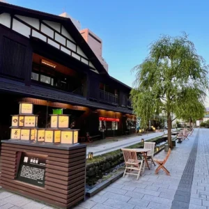 山形市内の七日町御殿堰。夕暮れの淡い青空の下、黒を基調とした和の趣ある街並みが水路に沿って続き、店頭に並ぶ温かな灯りをともした行灯風のオブジェと、水辺に置かれた木製のテーブルセットが日常から旅の情緒へと誘う風景。