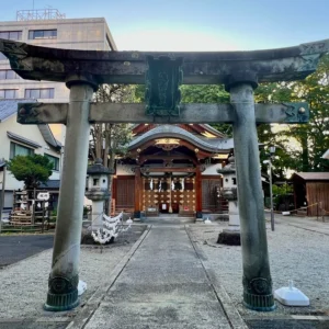 二年前の約束の地である、宿のすぐ目の前に佇む静かな神社の境内。使い込まれた石造りの鳥居が堂々とそびえ、奥に見える社殿の木造の扉や提灯、境内の砂利道が夕暮れの柔らかな光に包まれて奇跡的な再会の気配を漂わせている様子。