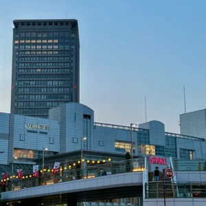 旅の余韻を残すJR山形駅の夕景。夕暮れの青く染まる空を背景に、高くそびえる高層ビルと駅舎の白い壁面、そして連絡通路に灯る温かな電球の明かりが、静けさと確かな未来への決意を感じさせる佇まい。