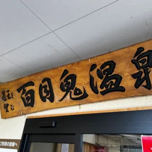 木製の茶色い板に力強い黒文字で「百目鬼温泉」と彫られた温泉の看板と、その下部の自動ドアや建物の一部。