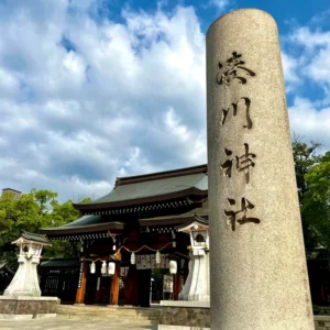 朝の澄んだ青空と柔らかな白い雲が広がる境内にて、重厚な石で造られた「湊川神社」と深く刻まれた巨大な社号標の石柱が垂直にそびえ立っている。その背後には伝統的な社殿の和風建築の屋根と、生命力に満ちた青々とした木々が美しく調和している。