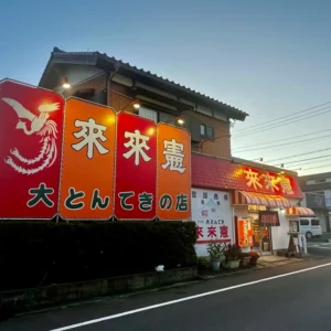 夕暮れ時の淡い光が広がるアスファルトの道路沿いに建つ、赤やオレンジ色の鮮やかな看板が目を引く建物の外観。「大とんてきの店」と力強い筆文字の看板が掲げられており、これから始まる長い道のりと旅の始まりを予感させる、旅の途中のワンシーンを捉えた一枚。