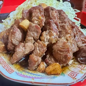 赤い模様が縁取られた中華風の平皿の上に、香ばしく焼き上げられ一口大に切り分けられた厚切りのとんてきが山のように高く積み上げられている。その隙間には大ぶりのニンニクの塊がゴロゴロと転がり、背後にはシャキシャキとした食感を思わせる山盛りの千切りキャベツが添えられた、圧倒的なボリューム感のある食事の様子。