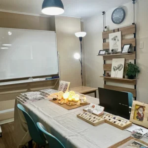 明るい光が差し込む室内に設営された鑑定会場（神戸駅A会場）の様子。壁側の大きなホワイトボードの前に、淡い麻のクロスがかけられた長机が置かれ、その上には温かみのあるランプの灯りや、美しく整えられた天然石の数々、ノートパソコンや資料が丁寧に並べてあり、これから始まる対話への準備が整えられた空間。