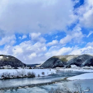 冬の澄んだ青空と大きく白い雲が広がるもと、白く雪化粧したなだらかな山々と川沿いの家並みが写っている。その手前では、冷涼な空気を映す青みがかった川の水が、岩肌を避けて絶えず流れている様子。