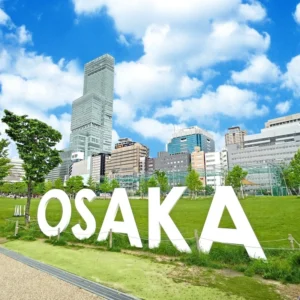 晴れ渡った青空に真っ白い雲が広がる日中の都市公園。緑豊かな芝生の手前には、立体的な「OSAKA」の巨大な白いモニュメントが堂々と配置され、その後方には高層ビル群や特徴的なタワー建築が幾重にも重なってそびえ立ち、現代都市の躍動感と開放的な空間が鮮やかに切り取られている。