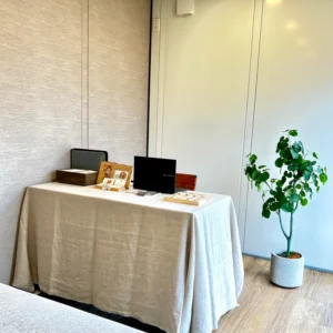 石屋蓮 高松鑑定会の会場内。明るいベージュのテーブルクロスが掛けられた鑑定用デスクが置かれ、その上にはパソコン、石を並べた木箱、店主の看板やパンフレットが整然と配置されています。デスクの右横には丸みのある葉が茂る鉢植えが置かれ、手前にはクライアント様用のソファの一部が見える、初めての方でも心穏やかに過ごせる落ち着いた鑑定空間の様子。